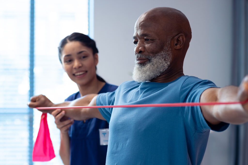 Smiling diverse female physiotherapist helping senior male patient exercise with band. Hospital, medical and healthcare services.