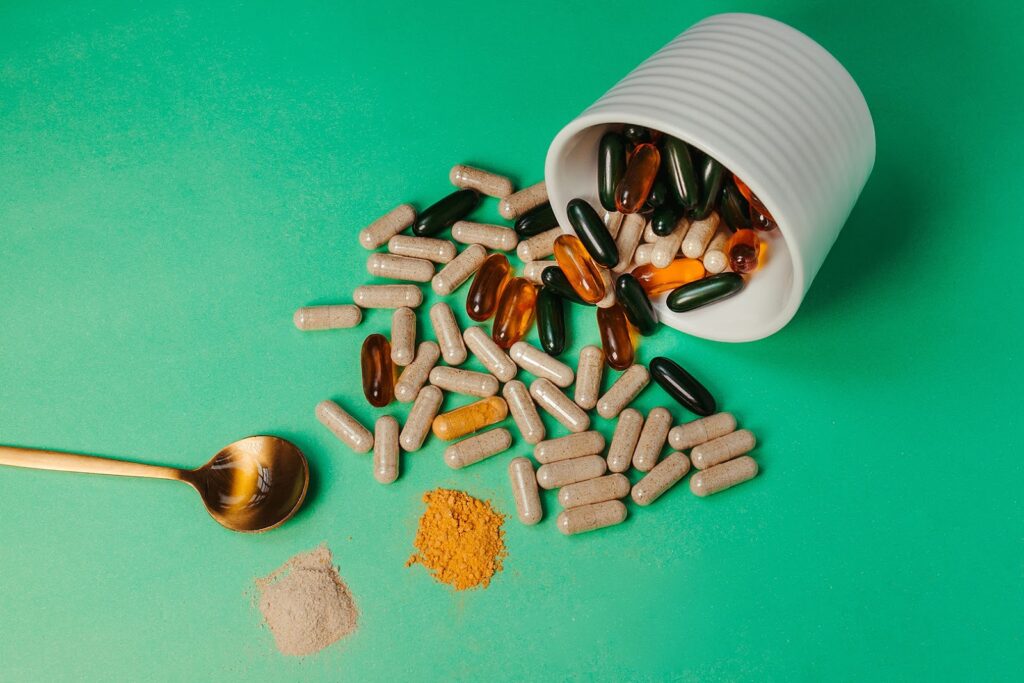 A green tabletop with an array of vitamin supplement capsules and pills and a spoon