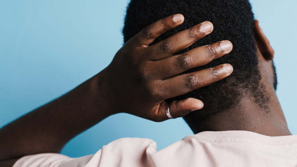 Close-up image of the back of a man's head that he holds with his hand while standing against a blue background.