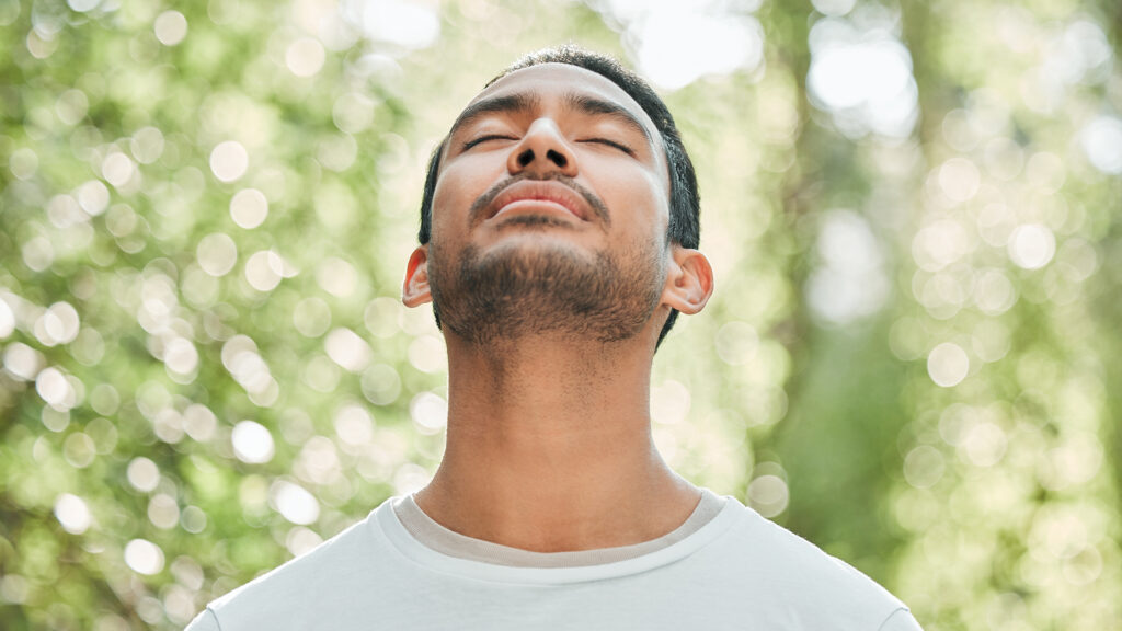 A man in the center of the image looks up with his eyes closed with trees in the background.