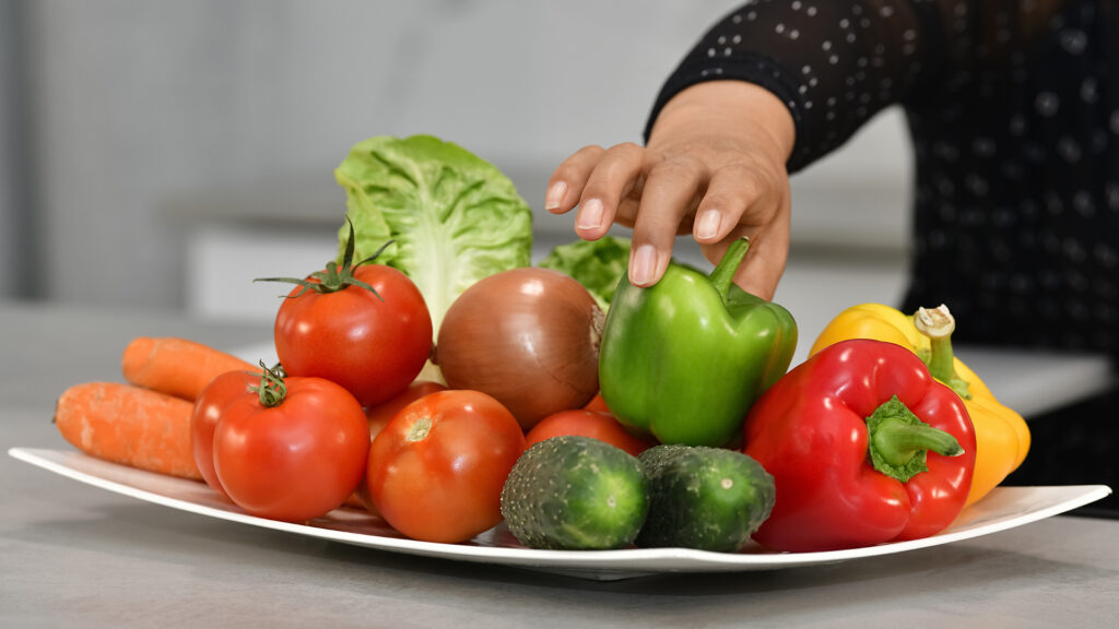 A woman's hand reaches to grab a green bell pepper that sits on a platter of various vegetables.