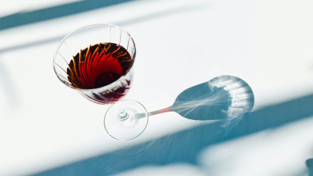 A glass full of a red alcoholic drink sits on a white table with dramatic blue shadows.