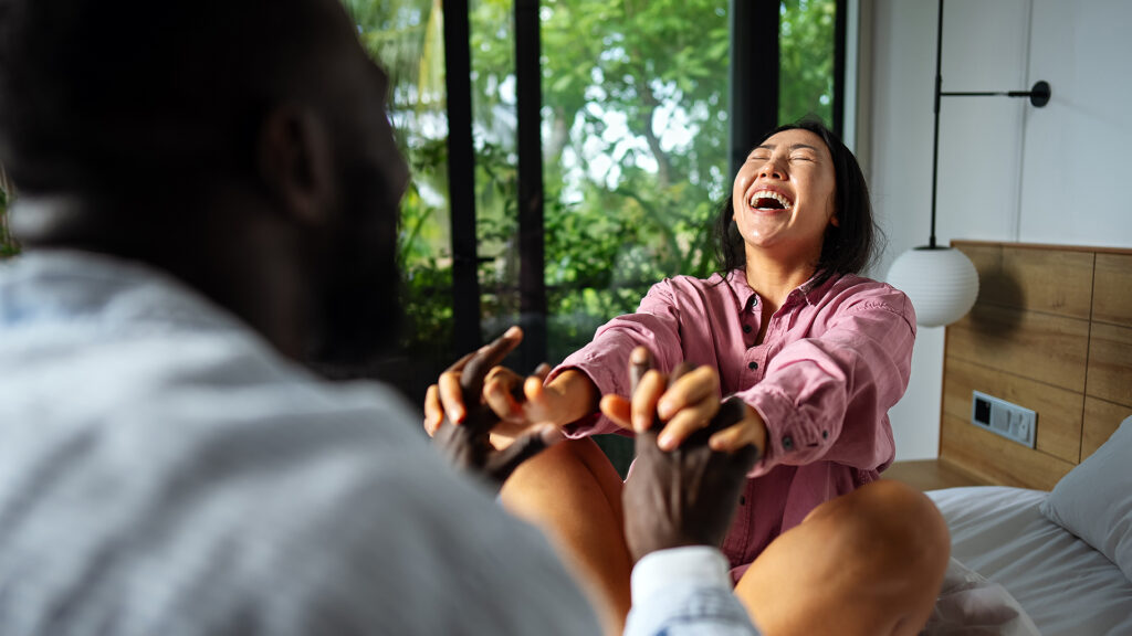 A laughing woman sitting on a bed holds hands with a man.