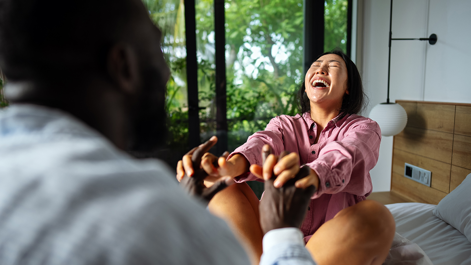 A laughing woman sitting on a bed holds hands with a man.