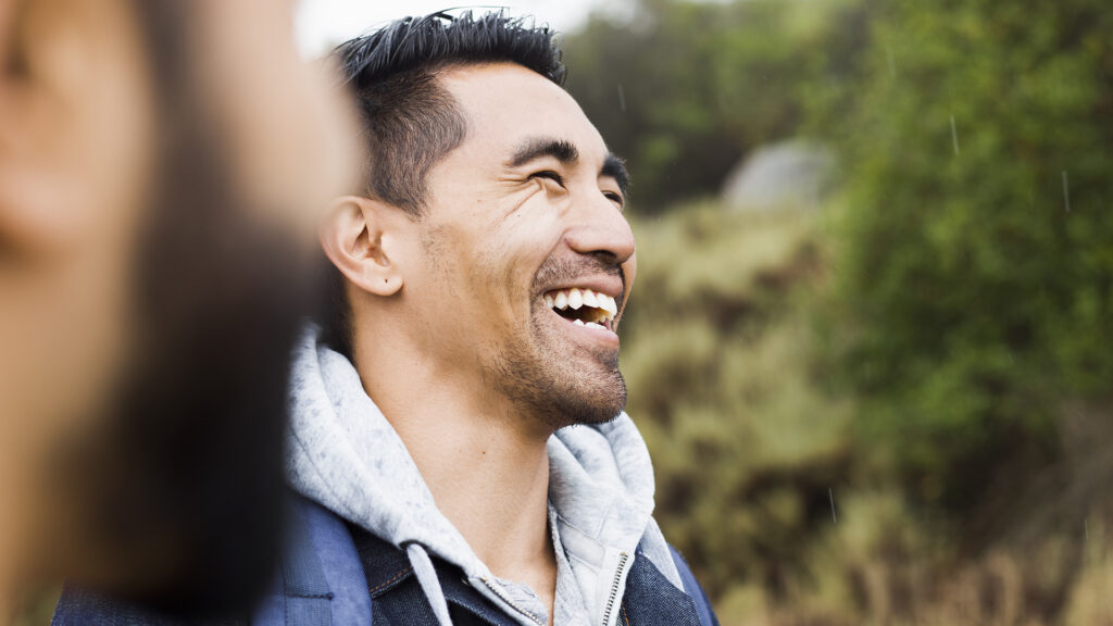 A man outdoors is laughing with the blurred face of another man is on the left side of the photo.