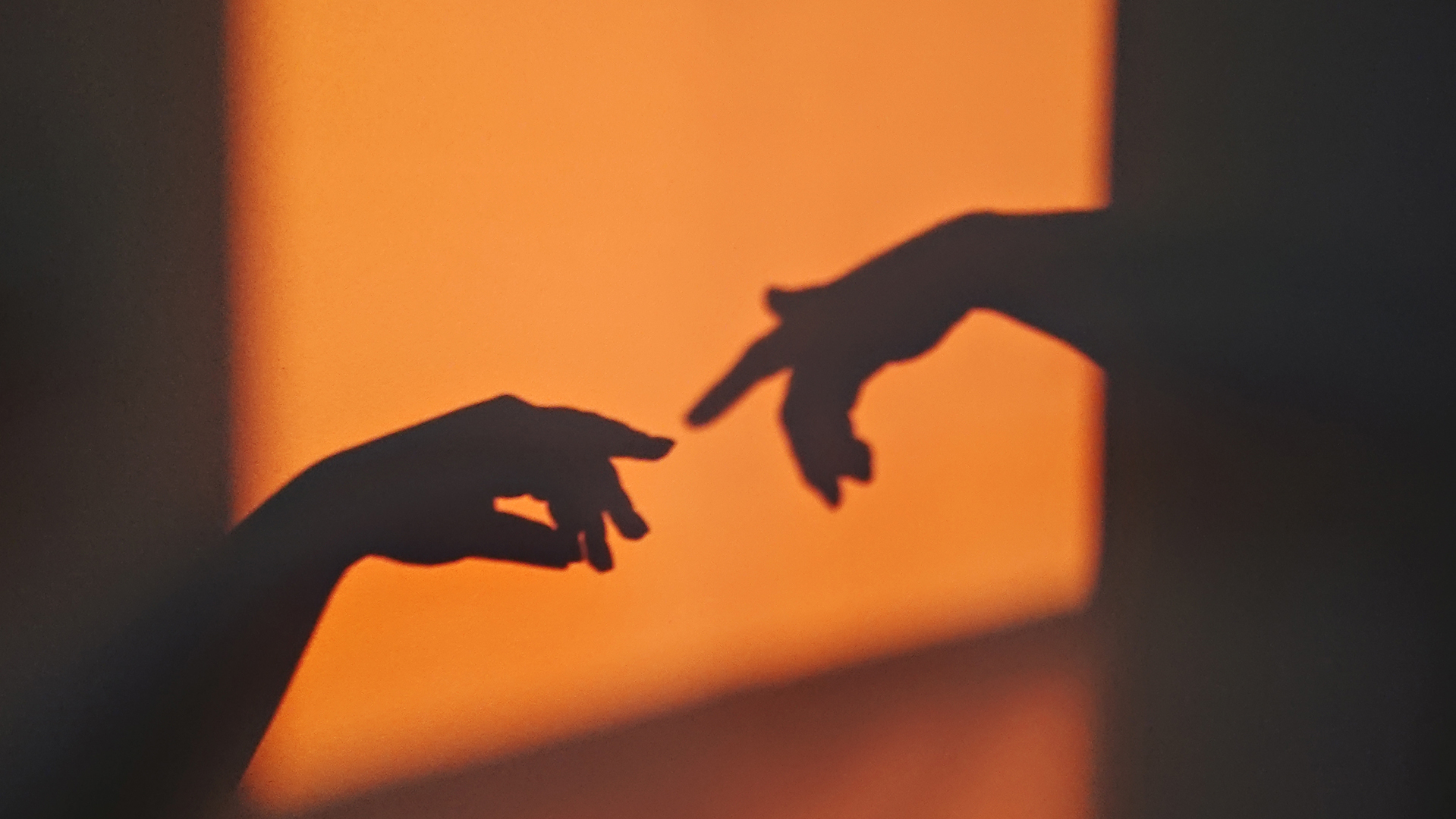 The shadows of two hands reach for each other against a wall lit up with an orange light.