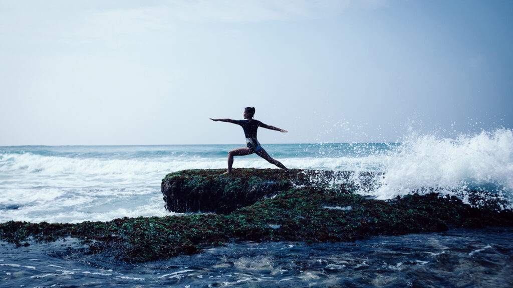 A woman does yoga on a large rock surrounded by crashing ocean waves.